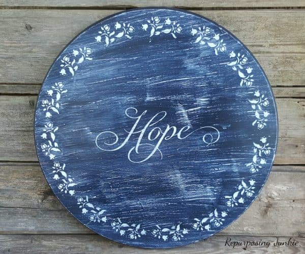 Round wooden sign painted in distressed navy blue with the word ‘Hope’ stenciled in elegant white script at the center, surrounded by a circular border of white floral stencil designs.