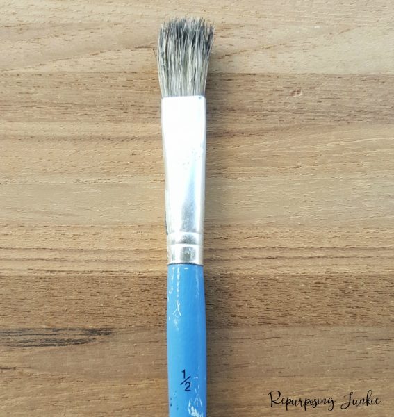 Close-up of a hand holding a stencil brush with a small amount of paint on the bristles, lightly tapping the brush onto a stencil placed over a wooden surface to demonstrate proper dry-brushing technique.
