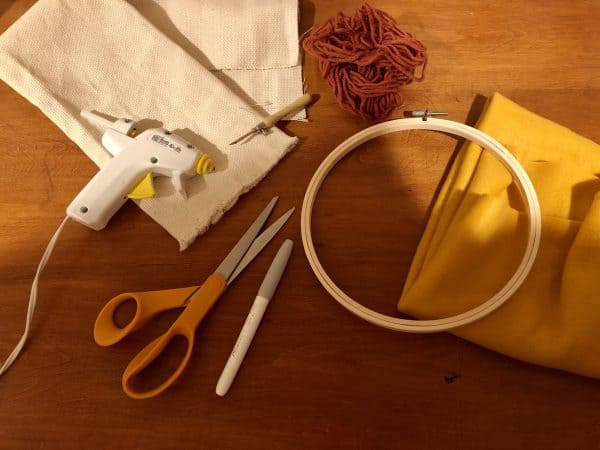 Supplies for adorable DIY phone pad made by a punch needle with acrylic yarn. Hot glue gun. Punch needle. Punch needle kit. Felt. Fleece. Fabric backing. Embroidery Hoop. Monks cloth. Fabric Scissors. Grey, Yellow, and White color scheme. Yarn crafts. Punch needle projects. Phone holder on desk. DIY phone holder.